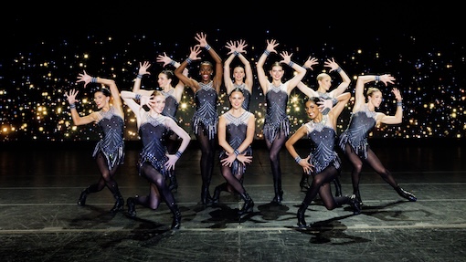 Rockettes in grey and black outfits, posing as a group on stage